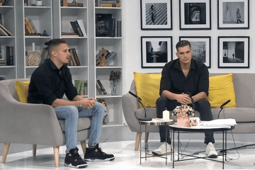 Two men in black shirts sit on a grey sofa with yellow pillows in a studio setting featuring bookshelves and framed wall art.