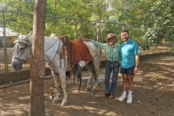 A man in a teal polo and an older man in a cowboy hat and flannel shirt stand together next to a saddled white horse in a fenced, sun-dappled yard.