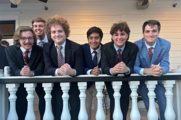 Six young men in formal suits and ties lean against a white balcony railing with classic balusters in front of a light-colored wooden house.