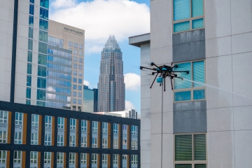 A professional-grade cleaning drone hovers beside a modern apartment building, spraying a stream of water onto the exterior windows against a city skyline featuring the Bank of America Corporate Center in Charlotte.