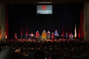 A wide view of a formal convocation ceremony on a stage with a "Davidson College" logo backdrop, featuring several faculty members in academic regalia seated around a student speaking at a podium