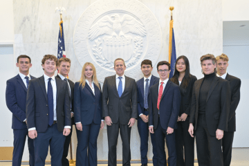 A group of ten professionally dressed students and a central official pose for a formal photo in front of a large, white architectural seal of the Federal Reserve Board of Governors.
