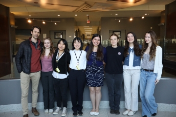 Seven young people stand side-by-side smiling in a brightly lit modern hallway with a decorative artistic ceiling.