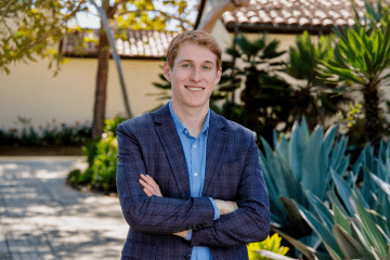 A young white man wearing a blue blazer in a tropical setting, arms crossed