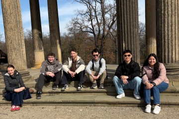 Six individuals sit smiling on the stone steps of a classical pavilion featuring large, fluted columns that frame a view of leafless trees under a clear blue sky.