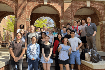 A large, diverse group of students and faculty pose together for a commemorative photo on the steps of a brick building with prominent arched entryways.