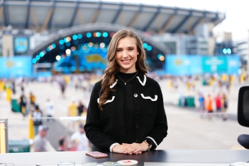 Courtney smiles at a desk with a stadium and large outdoor event stage blurred in the background.