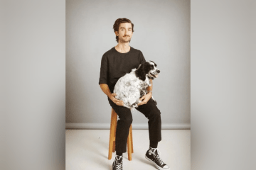 A man with a mustache sits on a wooden stool against a gray backdrop, wearing a black t-shirt, black pants, and black high-top sneakers while cradling a fluffy, black-and-white speckled dog on his lap.