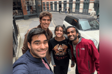 Four smiling friends take a high-angle selfie on a curved London street in front of a white compact car and a building labeled "Pub & Eating House."