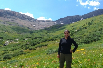 A smiling woman stands in a lush, green mountain valley filled with yellow and red wildflowers under a blue sky with light clouds.