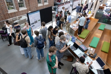 An overhead view of a busy atrium where students and faculty gather around research posters and technical demonstrations during a symposium.