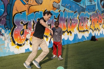 An adult man plays soccer with a young boy in front of a colorful graffiti mural