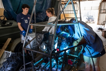 A student wearing a blue welding helmet and protective gloves welds a component onto the metal chassis of a custom off-road vehicle while two peers observe.