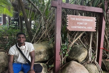 A young man sits on large rocks next to a wooden signpost reading "The Southernmost Point of Continental Asia," surrounded by tropical foliage.