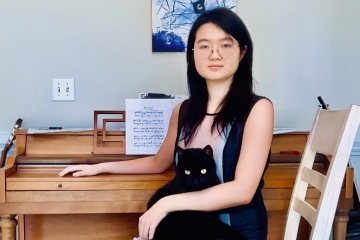 A female student with long black hair and glasses sits in front of a piano, holding a black cat