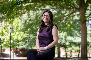 A smiling student wearing glasses and a dark purple sleeveless top sits outside on a stone ledge beneath leafy green trees.