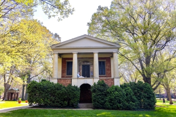 A person sits reading on the upper portico of a historic brick building with classical white columns, surrounded by leafy green trees and hedges on a sunny campus.