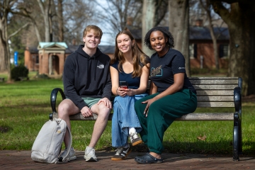 Three students sitting together on a wooden park bench, smiling at the camera.