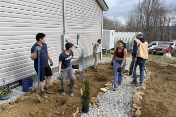 A group of volunteers works together on an outdoor landscaping project next to a house, digging trenches and laying gravel and stone borders.