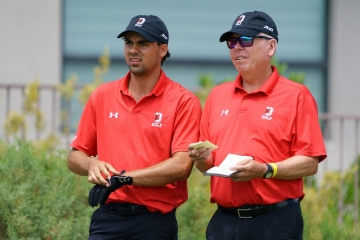 A young golfer adjusting a black glove next to a coach reviewing notes on a small yardage book. Both are wearing matching red Under Armour polo shirts and black caps branded with a white "D" and "GOLF" logo.