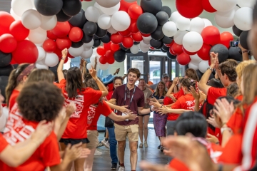 A young man walking down a hallway filled with red, white, and black balloons while crowd members in matching red shirts cheer and high-five him.