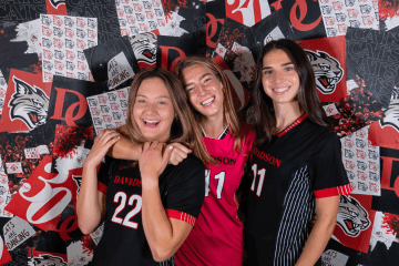 Three smiling female soccer players in Davidson College jerseys pose together in front of a red and black branded backdrop.
