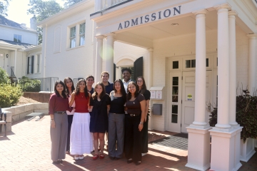 A diverse group of nine students smiling together for a group photo under the columned portico of a white "Admission" building.