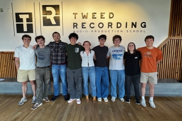 A group of eight young adults stands in a line with their arms around each other's shoulders, posing for a photo inside a modern room in front of a wall with a logo that reads "Tweed Recording Audio Production School."