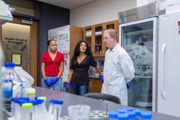 Dr. Thurtle-Schmidt, wearing a white lab coat, speaks with two student researchers wearing protective blue gloves inside a science laboratory.