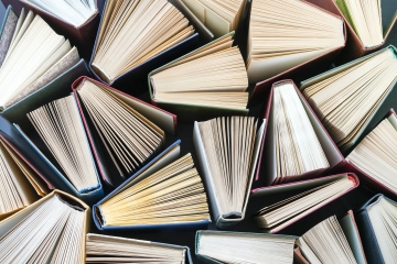 An overhead, top-down view of dozens of hardcover books arranged tightly together, each standing partially open with their paper pages fanning outward to form a geometric pattern.