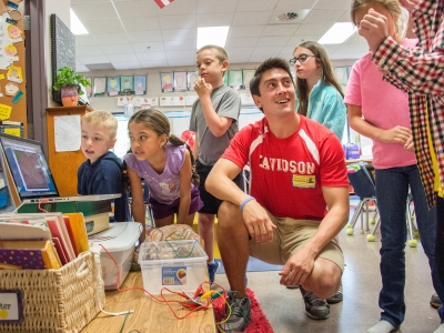 Davidson student wearing a Davidson soccer jersey stands amidst a group of young kids in their classroom and shows them math exercises on a laptop