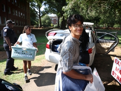 a young man moves into his dorm room while unpacking a car of stuff