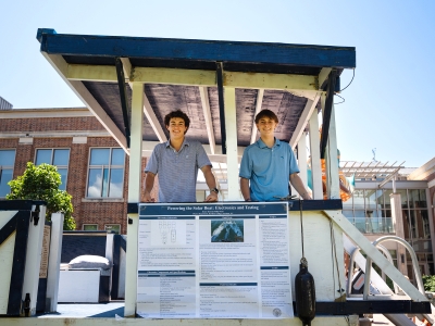 Two smiling male students in blue polo shirts stand proudly beside their solar boat project display titled "Powering the Solar Boat: Electronics and Testing," showcasing their engineering work under a small shelter with campus buildings visible in the background.