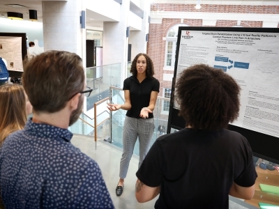 A student explains her research poster on virtual reality brain rehabilitation to a small group inside an academic building.