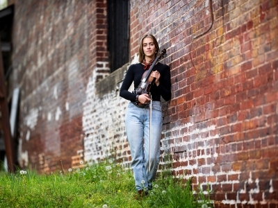 a young white woman standing against a brick wall holding a fiddle