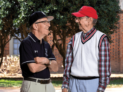 two older men stand together smiling looking at each other in front of a stadium, wearing Davidson College gear