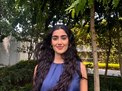 a young woman with long dark curly hair wearing a blue top standing outside in front of a set of green trees