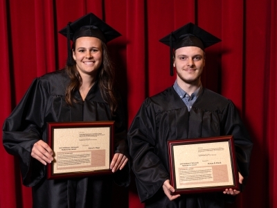 two college students in graduation regalia hold up framed awards