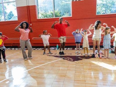 A group of diverse children and young adult leaders stand in a row on a gymnasium floor, smiling and flexing their muscles in a strong pose against a red and white wall.