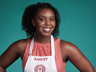 A smiling Black woman with curly hair wearing a white MasterChef apron with the name "ASHLEY" printed on it over a coral lace top, posed against a solid teal background.