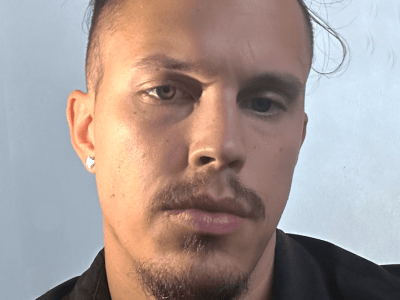 A man with a goatee, mustache, and dark hair pulled back looks directly into the camera while wearing a black collared shirt and a silver chain against a light-colored background.