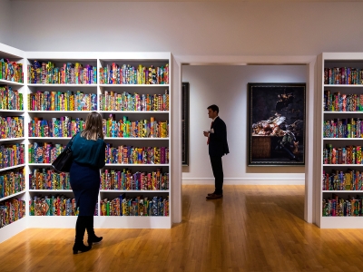 Visitors view an art installation featuring white bookshelves filled with books wrapped in vibrant, multicolored Dutch wax print fabrics.