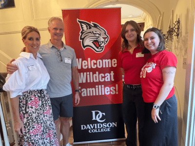 Two men and two women stand on a hardwood floor next to a red and black vertical banner that features a wildcat logo and the text, "Welcome Wildcat Families! Davidson College."