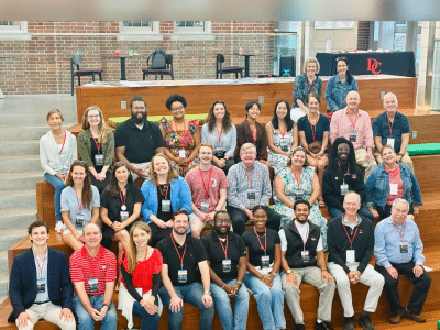 A diverse group of approximately twenty-five people sit on wide, tiered wooden bleachers indoors for a large group portrait.