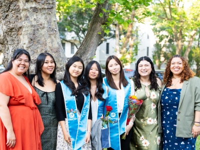 A group of seven smiling women pose outdoors in front of a large tree, including three graduates wearing light blue "College Track" sashes and holding flowers.
