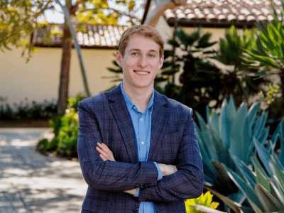 A young white man wearing a blue blazer in a tropical setting, arms crossed