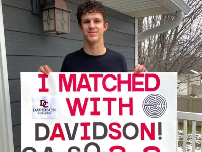A young man with curly hair stands outdoors on a deck holding a large white sign that reads "I MATCHED WITH DAVIDSON!" in bold red and black letters, featuring the Davidson College logo and a circular labyrinth icon.