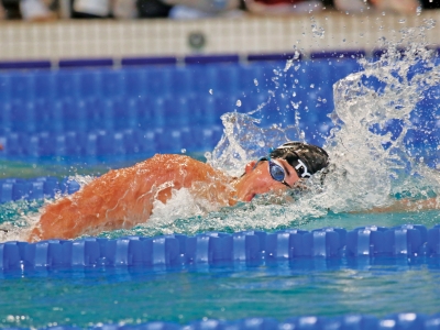 A swimmer wearing a black TYR swim cap and goggles breathes to the side while swimming freestyle in a pool lane, creating a large water splash.