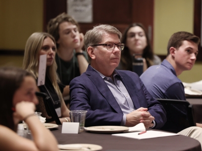 President Hicks with glasses and a blue blazer sits at a conference table with his hands clasped, listening intently alongside several young adults at a National Convening of Honor Councils event.