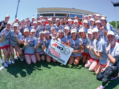 The Davidson College women's lacrosse team poses together on the field celebrating their victory, holding a sign that reads "Betterment A10 2026 Women's Lacrosse Champions" in front of the Davidson College Stadium.